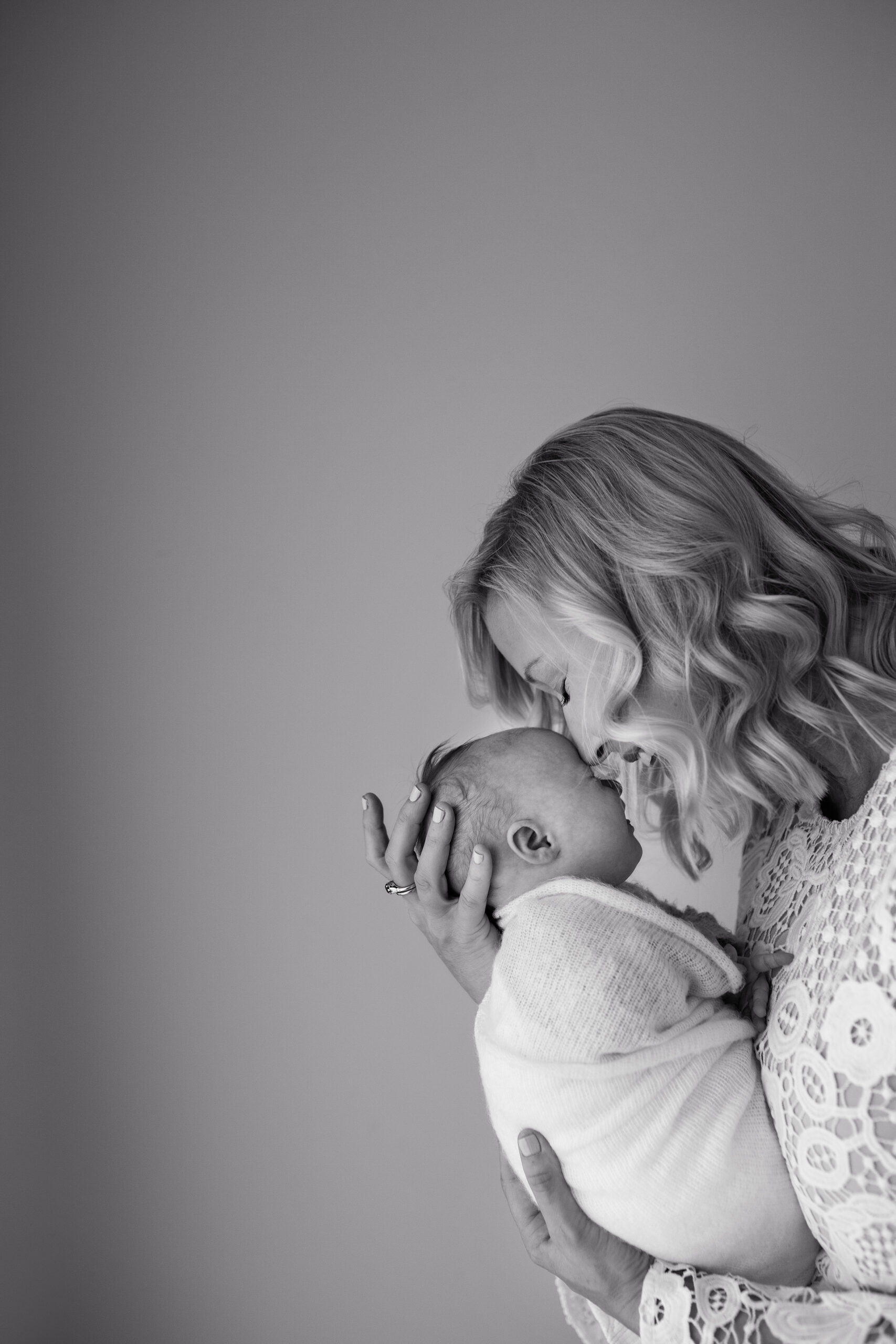 Black and white image of a mother with wavy hair holding her newborn baby. The mother's nose is touching her baby's forehead and they are both smiling.